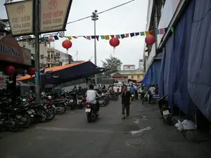 Bối cảnh là một con ngõ hoặc đường phố gần Chợ Đồng Xuân ở Hà Nội. Có các biển hiệu lớn treo trên cột ghi 'CHỢ ĐÊM ĐỒNG XUÂN' và chữ tiếng Anh 'NIGHT MARKET'. Phía dưới có một biển hiệu nhỏ hơn của một quầy hàng ghi 'Hải Sản Quầy số 11'. Con ngõ khá đông đúc, với rất nhiều xe máy đang đậu san sát nhau ở bên trái, có thể là một bãi giữ xe. Một số người đang đi lại trên đường, trong đó có một người đàn ông đang đi bộ và một người khác đang lái xe máy. Không gian được trang trí bằng một dây cờ nhiều màu và những chiếc đèn lồng đỏ treo ngang qua đường. Bên trái là các quầy hàng có mái che bằng bạt màu cam, trong khi bên phải là một tòa nhà được che phủ bởi các tấm bạt lớn màu xanh. Cảnh vật cho thấy một khu vực thương mại sầm uất, có thể là lối vào hoặc khu vực lân cận chợ.。（日本語訳）。背景はハノイのドンスアン市場近くの路地または通りです。柱には「CHỢ ĐÊM ĐỒNG XUÂN」と英語の「NIGHT MARKET」と書かれた大きな看板が掲げられています。その下には、「Hải Sản Quầy số 11」と書かれた屋台の小さな看板があります。路地はかなり混雑しており、左側にはたくさんのバイクが密集して停められています。おそらく駐輪場でしょう。通りには数人が行き来しており、その中には歩いている男性とバイクに乗っている人がいます。空間は色とりどりの旗の列と、通りを横切って吊るされた赤い提灯で飾られています。左側にはオレンジ色の防水シートで屋根が覆われた屋台があり、右側には大きな青い防水シートで覆われた建物があります。この光景は、市場の入り口またはその周辺地域である可能性のある、賑やかな商業エリアを示しています。