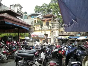 Bối cảnh là một khu vực sầm uất, có thể là một khu chợ hoặc bãi gửi xe ở Hà Nội. Tiền cảnh có rất nhiều xe máy đang được đỗ san sát nhau. Ở phía sau, nổi bật là một công trình kiến trúc cổ, một cổng tam quan sơn màu vàng với mái ngói cong, có thể là lối vào của một ngôi đền, chùa hoặc khu chợ truyền thống. Xung quanh là các tòa nhà dân sinh và cây xanh. Có nhiều hàng quán và cửa hiệu. Một quán ăn bên trái có biển hiệu lớn với các dòng chữ có thể đọc được một phần: '...UNG CHẢ CUA, MỰC LẨU...', 'NHẬN CÁ...', và 'CÁC MÓN NHẬU... BÒ...'. Ở phía xa hơn có các biển hiệu khác ghi 'QUÝ ANH'. Một vài chiếc ô che nắng được dựng lên, trong đó có một chiếc mang logo của thương hiệu nước 'Lavie'. Có người đang ngồi trong các hàng quán. Một tấm bạt lớn màu xanh lam bị rách che một phần góc trên bên phải của bức ảnh.。（日本語訳）。背景はハノイの活気のあるエリアで、市場か駐輪場かもしれない。前景にはたくさんのバイクがびっしりと駐車されている。後方には、古い建築物が際立っている。それは黄色に塗られ、反りのある瓦屋根を持つ三関門で、祠、寺、または伝統的な市場の入り口かもしれない。周りには民家や緑の木々がある。たくさんの飲食店や店がある。左側にある飲食店には大きな看板があり、部分的に読み取れる文字が書かれている：「...UNG CHẢ CUA, MỰC LẨU...」、「NHẬN CÁ...」、そして「CÁC MÓN NHẬU... BÒ...」。さらに遠くには「QUÝ ANH」と書かれた他の看板がある。いくつかの日よけの傘が立てられており、その中の一つにはミネラルウォーターブランド「Lavie」のロゴが入っている。飲食店の中には座っている人がいる。破れた大きな青い防水シートが、写真の右上の角の一部を覆っている。