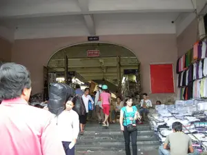 Bức ảnh chụp một lối vào của một khu chợ có mái che, đông đúc người qua lại. Phía trên lối vào hình vòm có một biển hiệu màu xanh ghi "CỬA SỐ 4". Bên trong vòm cửa, có một biển báo màu đỏ ghi "CẤM HÚT THUỐC" và chữ tiếng Anh "No Smoking。". Khung cảnh cho thấy hoạt động buôn bán tấp nập. Một người phụ nữ đang vác một túi hàng lớn màu đen trên vai đi xuống các bậc thang. Ở phía bên phải, rất nhiều quần áo, chủ yếu là áo phông, được xếp thành chồng cao và treo trên tường để bán. Một người đàn ông đang ngồi trên bậc thềm gần các chồng quần áo, có thể là người bán hàng. Nhiều người khác đang đi lại, ra vào chợ. Phía bên phải của lối vào có một bảng thông báo lớn màu đỏ nhưng không thể đọc được nội dung.。（日本語訳）。写真は、多くの人が行き交う、屋根付きの市場のある入り口を写している。アーチ型の入り口の上には、「CỬA SỐ 4」と書かれた青い看板がある。アーチの内側には、「CẤM HÚT THUỐC」と英語の「No Smoking。」が書かれた赤い標識がある。その光景は、活気のある商売の様子を示している。一人の女性が肩に大きな黒い荷物袋を担いで階段を下りている。右側では、主にTシャツであるたくさんの衣類が、高く積み重ねられ、壁に掛けて販売されている。一人の男性が衣類の山の近くの階段に座っており、販売員かもしれない。他の多くの人々が市場を行き来し、出入りしている。入り口の右側には大きな赤い掲示板があるが、内容は読み取れない。