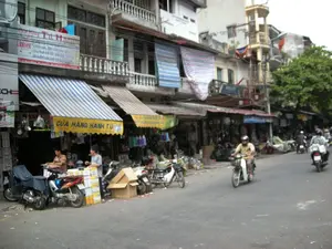 Bức ảnh chụp một con phố buôn bán sầm uất ở Việt Nam, có thể là Hà Nội. Dọc hai bên đường là những ngôi nhà ống nhiều tầng san sát nhau, với tầng trệt được sử dụng làm cửa hàng.。**Chữ viết trên các biển hiệu:**。*   Một biển hiệu trên mái hiên màu vàng có ghi rõ: "CỬA HÀNG HẠNH TÚ".。*   Phía trên, một tấm biển lớn hơn có các dòng chữ màu đỏ và xanh, một phần có thể đọc được là: "...TÚ HẠNH", "Chuyên cho thuê khách sạn, đầm cưới...", "Cho thuê bất động sản, đồ dùng...", "đã buffet các loại hàng...".。*   Một biển hiệu màu xanh ở phía xa có chữ "PHILIPS".。*   Có những biển hiệu khác nhưng chữ viết không thể đọc rõ.。**Bối cảnh và hoạt động:**。*   **Loại địa điểm:** Đây là một khu phố thương mại với nhiều cửa hàng bán lẻ ven đường.。*   **Hoạt động:** Nhiều người đang di chuyển trên đường bằng xe máy. Hoạt động mua bán đang diễn ra tại các cửa hàng. Trước "Cửa hàng Hạnh Tú", hai người đàn ông dường như đang sắp xếp hàng hóa hoặc giao dịch. Các cửa hàng trưng bày nhiều loại sản phẩm, có vẻ là đồ gia dụng và kim khí.。*   **Vật thể:** Có rất nhiều xe máy (xe gắn máy) đang đậu trên vỉa hè và lưu thông trên đường. Các mái hiên có sọc xanh-trắng và vàng được căng ra trước các cửa hàng. Nhiều thùng carton được xếp chồng lên nhau trên vỉa hè. Ở phía xa, có thể nhìn thấy một lá cờ Việt Nam (cờ đỏ sao vàng) treo trên một tòa nhà.。（日本語訳）。写真はベトナムの活気ある商店街を写したもので、おそらくハノイだと思われます。道の両側には多層階のチューブハウスが密集しており、1階は店舗として利用されています。**看板の文字：**。*   黄色い日よけの上にある看板には、「CỬA HÀNG HẠNH TÚ」（ハン・トゥ店）とはっきりと書かれています。*   その上には、より大きな看板があり、赤と青の文字の行があります。一部読み取れるのは、「...TÚ HẠNH」、「ホテル、ウェディングドレスのレンタル専門...」、「不動産、備品のレンタル...」、「...あらゆる種類の商品をビュッフェ...」です。*   遠くにある青い看板には「PHILIPS」という文字があります。*   他にも看板はありますが、文字ははっきりと読み取れません。**背景と活動：**。*   **場所の種類：**ここは道端に多くの小売店が並ぶ商業地区です。*   **活動：**多くの人々がバイクで道路を移動しています。店では売買活動が行われています。「Cửa hàng Hạnh Tú」の前では、2人の男性が商品を整理しているか、取引をしているように見えます。各店舗には多くの種類の商品が陳列されており、家庭用品や金物であるようです。*   **物体：**非常に多くのバイク（原動機付自転車）が歩道に駐車されたり、道路を通行したりしています。青と白の縞模様や黄色の縞模様の日よけが店の前に張られています。多くの段ボール箱が歩道に積み重ねられています。遠くには、建物に掲げられたベトナムの国旗（金星紅旗）が見えます。
