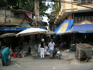 Bức ảnh chụp một con phố buôn bán sầm uất, có thể là một khu chợ ở Hà Nội. Các cửa hàng mặt tiền bày bán hàng hóa tràn ra vỉa hè.。Chữ viết trên các biển hiệu có thể nhận dạng được bao gồm tên các cửa hàng: "QUY ANH", "HƯƠNG HÀ" (bị che một phần), và "THANH THÂN".。Khung cảnh mô tả hoạt động kinh doanh trên một con phố. Các cửa hàng này chuyên bán đồ gia dụng, đặc biệt là các mặt hàng kim khí và thủy tinh như nồi, chảo, khay, ly, cốc được xếp chồng lên nhau. Hai người phụ nữ đang đứng giữa đường xem hàng, một người đội mũ bảo hiểm màu trắng. Bên cạnh họ là một chiếc xe máy đang đỗ. Ở phía bên trái, một chiếc xe máy khác được phủ bạt màu xanh lá cây. Phía bên phải có một chiếc xe đẩy rác bằng kim loại. Các cửa hàng sử dụng mái hiên màu xanh và những chiếc ô lớn để che nắng. Các tòa nhà là kiểu nhà ống đặc trưng của đô thị Việt Nam, với hệ thống dây điện chằng chịt ở phía trên.。（日本語訳）。写真は、ハノイのある市場と思われる、賑やかな商店街を写しています。店先では商品が歩道にまで溢れるように陳列されています。識別できる看板の文字には、「QUY ANH」、「HƯƠNG HÀ」（一部が隠れている）、そして「THANH THÂN」といった店名が含まれています。この光景は、ある通りでの商売の様子を描写しています。これらの店は家庭用品を専門に販売しており、特に鍋、フライパン、トレイ、グラス、カップといった金属製品やガラス製品が積み重ねられています。二人の女性が道の真ん中に立って商品を見ており、そのうちの一人は白いヘルメットをかぶっています。彼女たちの隣には、一台のバイクが停まっています。左側には、別のバイクが緑色のシートで覆われています。右側には、金属製のゴミ収集用の手押し車があります。店は日差しを遮るために、青い日よけや大きなパラソルを使用しています。建物はベトナムの都市に特徴的なチューブハウス（間口が狭く奥行きが深い家）の様式で、上部には電線が複雑に張り巡らされています。