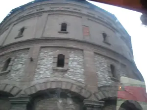 Bức ảnh chụp cận cảnh một công trình kiến trúc cổ hình trụ tròn, có khả năng là Tháp nước Hàng Đậu ở Hà Nội. Tòa tháp có hai tầng kiến trúc khác nhau. Tầng dưới được xây bằng đá hộc với các cửa sổ hình vòm. Tầng trên được trát vữa, có các cửa sổ nhỏ và hẹp hơn. Công trình mang dấu ấn của thời gian, với một số mảng tường bị bong tróc để lộ lớp gạch bên trong. Bức ảnh có vẻ được chụp từ bên trong một phương tiện đang di chuyển, vì hình ảnh hơi mờ và có một phần phản chiếu ở góc dưới bên phải. Không có chữ viết hay hoạt động của con người được ghi lại trong ảnh.。（日本語訳）。ハノイのハンザウ給水塔と思われる、円筒形の古い建築物のクローズアップ写真。この塔は2つの異なる建築様式の階層を持っている。下層階は粗石で造られており、アーチ型の窓がある。上層階は漆喰で塗られており、より小さく狭い窓がある。この建造物には時の流れが刻まれており、壁の一部が剥がれ落ちて中のレンガ層が露出している。写真は移動中の乗り物の中から撮影されたようで、画像が少しぼやけており、右下隅に一部反射が見られる。写真には文字や人間の活動は記録されていない。