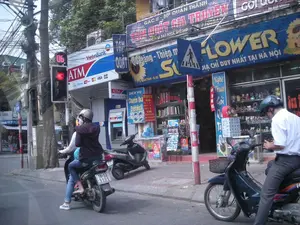 Bối cảnh là một con phố ở Hà Nội vào ban ngày. Trên đường, có hai người đang ngồi trên một chiếc xe máy, dường như đang dừng chờ đèn tín hiệu giao thông. Cả hai người đều đội mũ bảo hiểm. Ở bên phải, một người đàn ông khác cũng đang ngồi trên xe máy. Dọc vỉa hè là các cửa hàng san sát nhau. Có thể thấy rõ một trạm ATM của ngân hàng VietinBank. Bên cạnh đó là một cửa hàng lớn với biển hiệu màu xanh dương ghi chữ 'FLOWER' và 'ĐỊA CHỈ DUY NHẤT TẠI HÀ NỘI'. Các biển hiệu nhỏ hơn cho thấy cửa hàng này cung cấp các dịch vụ như 'Quà tặng - Thiệp mừng', 'DỊCH VỤ Chuyển Quà Điện Hoa THEO YÊU CẦU', và 'Nghệ Thuật Gói Quà'. Phía trên các cửa hàng là một biển hiệu khác ghi địa chỉ 'GÁC II - 80 QUÁN THÁNH' cùng với các dòng chữ '... GIA TRUYỀN' và 'PHỤC VỤ TẠI NHÀ RIÊNG'. Một cột đèn tín hiệu giao thông đang hiển thị đèn đỏ và số '06'. Ngoài ra còn có một biển báo giao thông cho người đi bộ sang đường và hệ thống dây điện chằng chịt đặc trưng.。（日本語訳）。背景はハノイのある昼間の通りです。路上には、バイクに二人乗りしている人がおり、信号待ちで停車しているようです。二人ともヘルメットをかぶっています。右側には、別の男性もバイクに乗っています。歩道沿いには店が軒を連ねています。VietinBankのATMがはっきりと見えます。その隣には、「FLOWER」と「ĐỊA CHỈ DUY NHẤT TẠI HÀ NỘI」（ハノイで唯一の住所）と書かれた青い看板を掲げた大きな店があります。より小さな看板には、この店が「Quà tặng - Thiệp mừng」（ギフト - グリーティングカード）、「DỊCH VỤ Chuyển Quà Điện Hoa THEO YÊU CẦU」（ご要望に応じたフラワー電報・ギフト配達サービス）、そして「Nghệ Thuật Gói Quà」（ギフトラッピングアート）などのサービスを提供していることが示されています。店の上には、「GÁC II - 80 QUÁN THÁNH」という住所と、「... GIA TRUYỀN」（...家伝）、「PHỤC VỤ TẠI NHÀ RIÊNG」（ご自宅でサービス提供）という文言が書かれた別の看板があります。信号機は赤信号と「06」という数字を表示しています。その他にも、歩行者横断用の交通標識と、特徴的な絡み合った電線システムがあります。