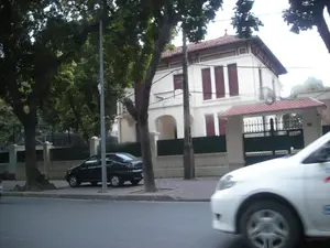 Bức ảnh chụp một cảnh đường phố ở Hà Nội. Nổi bật trong ảnh là một biệt thự hai tầng lớn mang phong cách kiến trúc kiểu Pháp, có tường sơn trắng, mái ngói đỏ và cửa sổ chớp màu đỏ. Biệt thự có ban công và được bao quanh bởi hàng rào và một cổng sắt lớn. Dọc vỉa hè có nhiều cây cổ thụ xanh tốt. Một chiếc ô tô màu đen đang đỗ trên vỉa hè gần các gốc cây. Ở phía trước bên phải, một phần của chiếc ô tô màu trắng đang di chuyển trên đường, hình ảnh bị nhòe do tốc độ. Không có người nào được nhìn thấy rõ trong ảnh. Trên cột cổng có một con số, có thể là số nhà, nhưng không thể đọc được. Chữ viết trên chiếc xe màu trắng cũng bị mờ và không thể nhận dạng.。（日本語訳）。ハノイのある街角の風景を撮った写真です。写真の中で際立っているのは、フランス風建築様式の大きな2階建てのヴィラで、白い壁、赤い瓦屋根、赤い鎧戸の窓があります。ヴィラにはバルコニーがあり、フェンスと大きな鉄の門で囲まれています。歩道に沿って、青々と茂る多くの古い木があります。木の根元近くの歩道に黒い車が1台停まっています。右手前では、白い車の一部が道路を移動しており、その画像は速度のためにぼやけています。門柱には数字があり、それは番地かもしれませんが、読み取れません。白い車に書かれた文字もぼやけていて、識別できません。