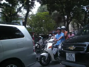 Cảnh tượng giao thông trên một con đường phố ở Hà Nội, có nhiều cây xanh ở hai bên. Dòng xe cộ đông đúc bao gồm cả ô tô và xe máy. Ở bên trái là một chiếc ô tô màu bạc, và ở phía trước bên phải là một chiếc ô tô màu đen. Nhiều người đang điều khiển xe máy giữa dòng xe cộ. Tất cả những người đi xe máy đều đội mũ bảo hiểm. Nổi bật là một người đàn ông mặc áo polo màu xanh lam đang lái một chiếc xe tay ga màu trắng, phía sau là một người phụ nữ mặc áo màu cam. Cảnh này mô tả hoạt động đi lại thường ngày trong thành phố.。（日本語訳）。ハノイのある街路の交通風景で、両側には緑の木々がたくさんあります。混雑した車両の流れには、自動車とバイクの両方が含まれています。左側には銀色の自動車が1台、そして右前方には黒色の自動車が1台あります。多くの人々が車両の流れの中でバイクを運転しています。バイクに乗っている人々は全員ヘルメットを着用しています。目立っているのは、青いポロシャツを着て白いスクーターを運転している男性で、その後ろにはオレンジ色の服を着た女性がいます。この光景は、都市における日常の移動の様子を描写しています。