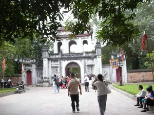 Bối cảnh là một khu di tích lịch sử tại Hà Nội, có khả năng cao là Văn Miếu - Quốc Tử Giám. Trong ảnh là một cổng tam quan lớn, cổ kính với ba lối vào hình vòm, là lối đi chính vào khu di tích. Nhiều người, dường như là du khách, đang đi bộ trên con đường lát gạch để vào tham quan. Một số người đang đi qua cổng, trong khi những người khác ngồi nghỉ trên bệ đá ven đường. Ở phía bên trái, có một vài chiếc xe máy đang đỗ. Hai bên lối đi được cắm nhiều lá cờ lễ hội truyền thống với màu sắc rực rỡ. Cây cối xanh tươi bao quanh, tạo bóng mát cho con đường. Trên cổng phụ bên trái có thể nhận ra hai chữ Hán "文廟" (Văn Miếu). Trên các cột của cổng chính cũng có các dòng chữ Hán Nôm được khắc dọc nhưng không thể đọc rõ do khoảng cách và chất lượng ảnh.。（日本語訳）。背景はハノイにある歴史的遺跡地区で、文廟（ヴァンミョウ）- 国子監（クオックトゥザム）である可能性が高いです。写真には、3つのアーチ型の入口を持つ、大きくて古風な三門（タムクアン）が写っており、これが遺跡地区への正門です。多くの人々、おそらく観光客が、見学のために入場しようとレンガ敷きの道を歩いています。何人かは門を通り抜けている一方で、他の人々は道端の石の台座に座って休んでいます。左側には、数台のバイクが停まっています。通路の両側には、色鮮やかな伝統的な祭りの旗が数多く立てられています。青々とした木々が周りを囲み、道に木陰を作っています。左側の副門には、「文廟」（Văn Miếu）という二つの漢字が認識できます。