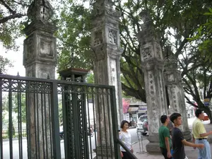 Bức ảnh chụp cổng vào của một di tích lịch sử ở Hà Nội. Nổi bật trong ảnh là bốn cột trụ bằng đá lớn, cổ kính, xếp thành hàng. Trên thân các cột trụ có chạm khắc hoa văn và các dòng chữ Hán Nôm được viết theo chiều dọc, tuy nhiên các chữ đã mờ và khó đọc rõ. Phía trước là một hàng rào và cổng lớn bằng sắt, sơn màu sẫm. Xung quanh có nhiều cây cổ thụ lớn, tán lá xum xuê. Ở phía bên phải, trên vỉa hè, có một nhóm người đang đi bộ, bao gồm một vài thanh niên và một người phụ nữ. Phía sau các cột trụ là khung cảnh đường phố với một chiếc ô tô đang đỗ và một vài người đi xe máy. Khung cảnh thể hiện sự hòa quyện giữa một công trình kiến trúc cổ và cuộc sống thường nhật.。（日本語訳）。ハノイのある歴史的遺跡の入り口を撮影した写真です。写真の中で目立っているのは、一列に並んだ4本の大きな石造りの古風な柱です。柱の本体には模様と縦書きのチュハンノム（漢字・チュノム）が彫られていますが、文字は薄れていてはっきりと読み取るのは困難です。手前には、濃い色に塗られた鉄製の大きな柵と門があります。周りには、葉が青々と茂った大きな古木がたくさんあります。右側の歩道には、数人の若者と一人の女性を含む、歩いている人々のグループがいます。柱の後ろには、一台の駐車中の自動車と数人のバイクに乗った人々がいる街の風景が広がっています。この光景は、古い建築物と日常生活との調和を表しています。