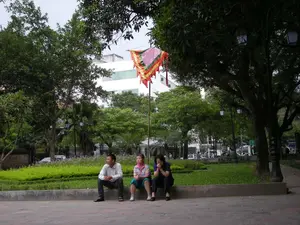 Bối cảnh là một công viên hoặc không gian công cộng ở Hà Nội. Ba người, gồm một người đàn ông và hai người phụ nữ, đang ngồi nghỉ ngơi trên một bờ tường bê tông thấp ven lối đi lát gạch. Người đàn ông mặc áo sơ mi sáng màu, hai người phụ nữ ngồi bên cạnh ông. Họ dường như đang thư giãn và ngắm cảnh. Xung quanh có rất nhiều cây xanh lớn, tán lá rậm rạp tạo bóng mát. Ở trung tâm bức ảnh, có một cột cờ treo một lá cờ hội truyền thống hình tam giác, nhiều màu sắc rực rỡ (hồng, vàng, xanh lá). Phía sau họ là một thảm cỏ xanh, các luống hoa và một hàng rào sắt. Xa hơn trong hậu cảnh có thể thấy một tòa nhà hiện đại màu trắng. Dọc theo công viên có những cột đèn đường mang phong cách cổ điển. Không có chữ viết nào có thể đọc được trong ảnh.。（日本語訳）。背景はハノイのある公園または公共空間です。一人の男性と二人の女性を含む3人が、煉瓦敷きの通路のそばにある低いコンクリートの壁の縁に座って休んでいます。男性は明るい色のシャツを着ており、二人の女性が彼の隣に座っています。彼らはリラックスして景色を眺めているようです。周りには大きな緑の木がたくさんあり、鬱蒼と茂った葉が日陰を作っています。写真の中央には、一本の旗竿があり、鮮やかな多色（ピンク、黄色、緑）の三角形の伝統的な祭りの旗が掲げられています。彼らの後ろには、一面の緑の芝生、花壇、そして鉄の柵があります。さらに奥の背景には、白い近代的な建物が見えます。公園に沿って、クラシックなスタイルの街灯があります。写真の中には、読み取れる文字はありません。