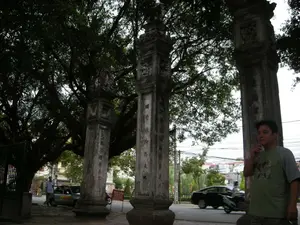 Bối cảnh là một không gian ngoài trời, có vẻ như là cổng vào của một di tích lịch sử hoặc tôn giáo như đình, chùa ở Hà Nội. Nổi bật là các cột trụ bằng đá lớn, cũ kỹ, có thể là một phần của cổng tam quan. Trên các cột trụ có chạm khắc hoa văn và các dòng chữ Hán hoặc chữ Nôm chạy dọc thân cột, tuy nhiên chữ viết đã mờ và khó đọc. Phía trên là một cây cổ thụ rất lớn, tán lá xum xuê che bóng mát. Một người đàn ông mặc áo phông màu xanh đang đứng ở phía trước bên phải, cạnh một cột đá. Phía sau, trên con đường nhựa, có các phương tiện giao thông như ô tô và xe máy đang di chuyển. Có một biển báo màu đỏ với chữ 'YÊU CẦU' nhưng phần nội dung còn lại không rõ. Cảnh vật cho thấy sự giao thoa giữa một không gian cổ kính, trang nghiêm và nhịp sống đô thị hiện đại.。（日本語訳）。背景は屋外の空間で、ハノイにある亭や寺院のような歴史的または宗教的な遺跡の入り口のようです。目立っているのは、大きくて古い石柱で、三関門（タムクアン）の一部かもしれません。柱には文様と、柱身に沿って縦に書かれた漢文またはチュノムの文字列が彫られていますが、文字はかすれていて読みにくいです。上方には非常に大きな古木があり、葉が青々と茂って涼しい木陰を作っています。青いTシャツを着た男性が、前面右側の石柱の隣に立っています。後方では、アスファルトの道路上を自動車やバイクなどの乗り物が移動しています。「YÊU CẦU」（要求）という文字が書かれた赤い看板がありますが、残りの内容ははっきりしません。この光景は、古風で荘厳な空間と現代の都市生活のリズムとの交錯を示しています。