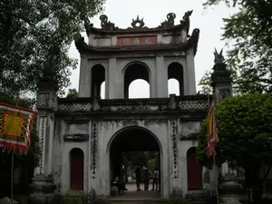 Bức ảnh chụp cổng chính của một khu di tích lịch sử, có kiến trúc cổ xưa. Đây là cổng Văn Miếu Môn, lối vào của Văn Miếu - Quốc Tử Giám ở Hà Nội.。**Chữ viết:**。*   Trên tấm biển ở tầng hai của cổng có khắc ba chữ Hán lớn màu đỏ: 「文廟門」 (Văn Miếu Môn).。*   Trên các cột trụ ở hai bên cổng chính có khắc các dòng chữ Hán theo chiều dọc, nhưng không thể đọc rõ nội dung.。**Bối cảnh và hoạt động:**。*   **Địa điểm:** Cổng vào một khu di tích lịch sử và văn hóa (Văn Miếu Môn). Cổng có hai tầng với các lối đi hình vòm, mang vẻ cổ kính.。*   **Hoạt động:** Một nhóm người, có thể là du khách, đang đứng bên trong lối đi chính của cổng để tham quan.。*   **Vật thể:**。*   Cổng tam quan bằng đá.。*   Mái cổng cong và các chi tiết điêu khắc trang trí hình linh vật.。*   Các lá cờ hội truyền thống được cắm ở hai bên: một lá cờ vuông lớn màu vàng với viền xanh lá và đỏ, và một lá cờ lễ hội khác.。*   Cây cối xanh tươi, bao gồm cả cây cổ thụ, bao quanh công trình.。（日本語訳）。歴史的遺跡の正門を撮影した写真で、古い建築様式です。これはハノイにある文廟・國子監の入り口である文廟門です。**文字:**。*   門の2階にある扁額には、赤色の大きな漢字3文字「文廟門」（Văn Miếu Môn）が刻まれています。*   正門の両側にある柱には、縦書きの漢文が刻まれていますが、内容ははっきりと読み取れません。**背景と活動:**。*   **場所:** 歴史文化遺跡（文廟門）の入り口の門。門は2階建てで、アーチ型の通路があり、古風な趣があります。*   **活動:** 観光客と思われる一団が、見学のために門の主要な通路の内側に立っています。*   **物体:**。*   石造りの三関門。*   湾曲した門の屋根と、霊獣をかたどった装飾彫刻のディテール。*   両脇には伝統的な祭りの旗が立てられています。緑と赤の縁取りがある大きな黄色の四角い旗と、もう一つの祭りの旗です。*   古木を含む青々とした木々が、建物を囲んでいます。