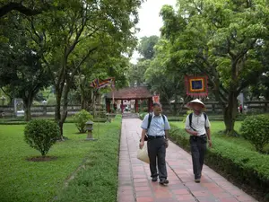 Bức ảnh chụp một khung cảnh thanh bình bên trong một khu di tích lịch sử, có khả năng là một ngôi đền hoặc chùa ở Hà Nội. Nơi đây có một khu vườn rộng lớn với nhiều cây cổ thụ xanh tươi, bãi cỏ được cắt tỉa cẩn thận và một lối đi bộ lát gạch thẳng tắp. Ở phía trước, hai người đàn ông, có vẻ là du khách, đang đi dạo tham quan trên lối đi. Người đàn ông bên phải đội một chiếc nón lá truyền thống, đeo ba lô. Người đàn ông bên trái mặc áo sơ mi xanh, cũng đeo ba lô và xách một chiếc túi đan lát. Ở phía sau là một công trình kiến trúc cổ với mái ngói và các cột sơn đỏ, có thể là cổng vào hoặc một gian nhà trong khu di tích. Dọc theo lối đi và trong vườn có treo những lá cờ hội nhiều màu sắc, tạo không khí trang trọng. Có thể nhìn thấy một chiếc đèn đá đặt trên bãi cỏ. Không có văn bản nào có thể đọc được rõ ràng trong ảnh.。（日本語訳）。写真は、ハノイにある寺院か仏塔と思われる史跡内の、穏やかな風景を捉えています。ここには、青々とした多くの古木、手入れの行き届いた芝生、そしてまっすぐに続くレンガ敷きの遊歩道がある広大な庭園があります。手前では、観光客と思われる2人の男性が遊歩道を散策しています。右側の男性は伝統的なノンラー（円錐形の葉笠）をかぶり、バックパックを背負っています。左側の男性は青いシャツを着て、同じくバックパックを背負い、編みかごのバッグを手に持っています。後方には、瓦屋根と赤く塗られた柱を持つ古い建築物があり、それは入口の門か、史跡内の建物である可能性があります。遊歩道沿いや庭園には色とりどりの祭りの旗が掲げられ、荘厳な雰囲気を醸し出しています。芝生の上には石灯籠が見えます。写真の中には、はっきりと読み取れる文字はありません。