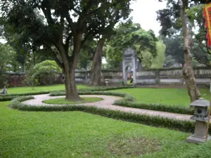 Bức ảnh chụp một không gian sân vườn hoặc công viên yên tĩnh, có khả năng là một phần của một khu di tích lịch sử ở Hà Nội. Nổi bật trong ảnh là một bãi cỏ xanh rộng lớn, được chăm sóc kỹ lưỡng. Ở trung tâm là một cây cổ thụ to lớn, được bao quanh bởi một lối đi lát gạch uốn lượn, viền bởi hàng cây bụi thấp. Ở phía sau, có một bức tường đá và một cổng vào mang phong cách kiến trúc cổ. Phía trước bên phải có một chiếc đèn đá. Một người đang ngồi ở phía xa bên trái. Cảnh vật mang lại cảm giác thanh bình, tĩnh lặng. Không có văn bản nào có thể đọc được.。（日本語訳）。写真は、静かな庭園または公園の空間を写しており、ハノイにある歴史的遺跡の一部である可能性があります。写真の中で目立っているのは、手入れの行き届いた広大な緑の芝生です。中央には一本の大きな古木があり、低木の生け垣で縁取られた湾曲したレンガ敷きの小道に囲まれています。後方には、石垣と古い建築様式の門があります。右手前には石灯籠が一つあります。左奥に一人の人が座っています。景色は、平和で静かな感覚をもたらします。読み取れる文字はありません。