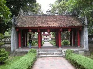 Bức ảnh chụp một công trình kiến trúc cổ của Việt Nam, có thể là một trong các cổng hoặc nhà bia trong một khu di tích lịch sử ở Hà Nội. Công trình có dạng một gian nhà mở với các cột trụ tròn bằng gỗ được sơn màu đỏ son. Mái được lợp bằng ngói âm dương màu nâu đỏ, đã nhuốm màu thời gian, trên nóc mái có các chi tiết điêu khắc trang trí. Dưới hiên nhà có treo những chiếc đèn lồng màu đỏ. Một lối đi lát gạch dẫn xuyên qua gian nhà, hai bên là các hàng cây cảnh được cắt tỉa gọn gàng. Một người phụ nữ, có lẽ là du khách, đang đứng bên trong gian nhà, tay cầm một chiếc túi xách màu đen. Nhìn xuyên qua cổng, ở phía xa có thể thấy một công trình cổng khác và một vài người đang đi lại. Không có chữ viết nào có thể đọc được trong ảnh.。（日本語訳）。写真はベトナムの古い建築物を写したもので、ハノイの歴史的遺跡地区にある門または石碑堂の一つである可能性があります。この建物は開かれた一間の家の形をしており、朱色に塗られた木製の丸い柱があります。屋根は赤褐色の陰陽瓦で葺かれ、時の色を帯びており、屋根の棟には装飾的な彫刻のディテールがあります。軒下には赤い提灯が吊るされています。レンガ敷きの通路がその一間を通り抜けており、両側にはきれいに刈り込まれた観賞用の木々が並んでいます。一人の女性、おそらく観光客が、その一間の中に立っており、黒いハンドバッグを手に持っています。門を通して見ると、遠くに別の門の建物と数人の人々が歩いているのが見えます。写真の中には読み取れる文字はありません。