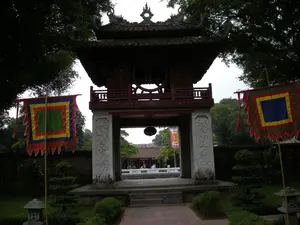 Bức ảnh chụp Gác Khuê Văn, một công trình kiến trúc cổ tại một khu di tích lịch sử, có khả năng là Văn Miếu - Quốc Tử Giám ở Hà Nội. Công trình này là một lầu gác hai tầng. Tầng trên được làm bằng gỗ sơn đỏ, có mái ngói cong đặc trưng của kiến trúc Á Đông và bên trong treo một quả chuông lớn. Tầng dưới được chống đỡ bởi bốn cột đá vuông lớn có chạm khắc hoa văn. Hai bên lối đi dẫn vào Gác Khuê Văn có cắm hai lá cờ hội ngũ sắc, là loại cờ truyền thống thường dùng trong các lễ hội ở Việt Nam. Xung quanh là không gian sân vườn với nhiều cây xanh, bãi cỏ và các đèn đá trang trí. Nhìn qua cổng vòm, có thể thấy một khoảng sân khác và một vài người dường như là du khách đang đi dạo.。（日本語訳）。ハノイの文廟・國子監と思われる歴史遺跡にある古い建築物、奎文閣（けいぶんかく）を写した写真。この建造物は二層の楼閣です。上層は赤く塗られた木で造られ、東アジア建築特有の反りのある瓦屋根を持ち、内部には大きな鐘が吊るされています。下層は、模様が彫刻された四本の大きな四角い石柱によって支えられています。奎文閣へと続く通路の両脇には、ベトナムの祭りでよく使われる伝統的な旗である五色旗が二本立てられています。周囲は多くの緑樹、芝生、そして装飾的な石灯籠がある庭園空間です。アーチ門を通して見ると、別の中庭と、散策している観光客と思われる数人の人々が見えます。