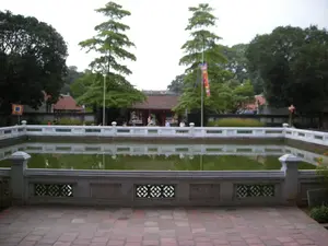 Bức ảnh chụp một khung cảnh bên trong một khu di tích lịch sử ở Hà Nội, có khả năng là Văn Miếu - Quốc Tử Giám. Ở vị trí trung tâm là một hồ nước lớn hình vuông, có thể là giếng Thiên Quang, với mặt nước tĩnh lặng màu xanh lục. Xung quanh hồ được bao bọc bởi lan can bằng đá hoặc xi măng sơn trắng, có các trụ và các ô hoa văn trang trí. Phía trước hồ là một khoảng sân rộng được lát bằng gạch nung màu đỏ. Ở phía sau hồ là một công trình kiến trúc cổ với mái ngói đỏ, hai bên lối vào có hai cây cao, thẳng đứng. Nhiều cây xanh tươi tốt khác mọc xung quanh, tạo nên một không gian yên tĩnh. Có một vài cột cờ được dựng lên, treo các lá cờ lễ hội, bao gồm một lá cờ phướn dài nhiều màu và một lá cờ vuông màu vàng viền đỏ. Ở phía xa, có thể thấy một vài người đang đi dạo tham quan khu di tích. Không có chữ viết nào có thể đọc được một cách rõ ràng.。（日本語訳）。ハノイの歴史的遺跡の内部の風景を撮影した写真で、おそらく文廟・國子監（ヴァンミュー・クオックトゥーザム）でしょう。中央には大きな四角い池があり、おそらく天光井（ティエンクアンの井戸）で、水面は静かで緑色をしています。池の周りは、石または白く塗られたセメント製の手すりで囲まれており、柱と装飾的な模様のパネルがあります。池の前には、赤レンガで舗装された広い中庭があります。池の後ろには、赤い瓦屋根の古い建築物があり、入り口の両側には2本の背の高い、まっすぐな木があります。周りには他にも多くの青々とした木々が生い茂り、静かな空間を作り出しています。いくつかの旗竿が立てられ、祭りの旗が掲げられています。それには、多色の長い幟（のぼり）と、赤で縁取られた黄色の四角い旗が含まれます。遠くには、遺跡を散策し見学している数人の人々が見えます。はっきりと読み取れる文字はありません。