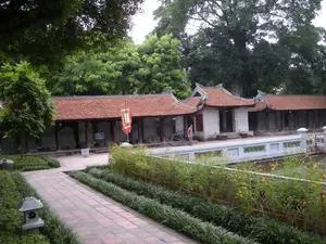 Bức ảnh chụp một khung cảnh bên trong một khu di tích lịch sử, có khả năng là Văn Miếu - Quốc Tử Giám ở Hà Nội. Nổi bật trong ảnh là một dãy nhà kiến trúc cổ với mái ngói đỏ và các cột gỗ, nơi trưng bày các tấm bia đá. Dưới mái hiên, nhiều tấm bia đá lớn (Bia Tiến sĩ) được đặt trang trọng trên lưng rùa đá. Phía trước dãy nhà bia là một hồ nước nhỏ được bao quanh bởi lan can đá có hoa văn. Lối đi trong khuôn viên được lát gạch đỏ, uốn lượn quanh những bồn hoa và thảm cỏ xanh. Ở phía trước, có những bụi hoa màu vàng cam và một chiếc đèn đá trang trí. Cảnh quan được bao phủ bởi nhiều cây xanh, bao gồm cả những cây cổ thụ, tạo nên một không gian thanh bình, cổ kính. Có một vài người trong ảnh, dường như là du khách đang tham quan; một người đang đứng và một người khác đang nghỉ ngơi trên bệ đá. Một lá cờ hội truyền thống được cắm gần đó. Có một tấm biển báo màu xanh nhưng không thể đọc được chữ. Các ký tự trên bia đá và tường nhà cũng không thể nhận dạng được do khoảng cách và độ phân giải.。（日本語訳）。写真は、ある史跡内の風景を写したもので、ハノイの文廟・國子監である可能性が高い。写真の中でひときわ目立つのは、赤い瓦屋根と木製の柱を持つ古い建築様式の建物が並んでいる様子で、そこには石碑が展示されている。石碑が並ぶ建物の前には、模様が施された石の手すりで囲まれた小さな池がある。敷地内の通路は赤いレンガで舗装され、花壇や緑の芝生の周りを曲がりくねっている。手前には、オレンジがかった黄色の花の茂みと装飾的な石灯籠がある。風景は古木を含む多くの緑の木々に覆われ、静かで古風な空間を作り出している。写真には数人の人物が写っており、観光客のようだ。一人は立っており、もう一人は石の台座で休んでいる。近くには伝統的な祭りの旗が立てられている。青い看板があるが、文字は読み取れない。石碑や建物の壁の文字も、距離と解像度のために識別できない。