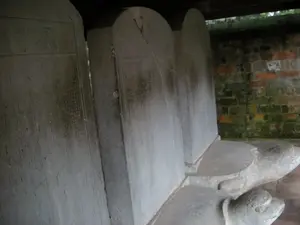 Bức ảnh chụp một dãy các bia đá được đặt trên lưng rùa đá tại một khu di tích lịch sử, có khả năng là Văn Miếu - Quốc Tử Giám ở Hà Nội. Các tấm bia này, được gọi là bia Tiến sĩ, được làm bằng đá, có hình chữ nhật với phần đỉnh được bo tròn. Trên bề mặt bia có khắc dày đặc các ký tự, có thể là chữ Hán, nhưng đã bị mờ và bào mòn theo thời gian nên không thể đọc rõ. Mỗi tấm bia được đặt trang trọng trên lưng một con rùa đá lớn, được điêu khắc tinh xảo. Phía sau là một bức tường gạch cũ đã bám rêu, và phía trên là một phần của mái hiên bằng gỗ, cho thấy các bia đá này được bảo quản trong một công trình kiến trúc cổ. Khung cảnh tĩnh lặng, không có hoạt động của con người, chỉ có các hiện vật lịch sử.。（日本語訳）。写真は、歴史的遺跡、おそらくハノイの文廟・國子監で、石亀の背中に乗せられた石碑の列を写している。これらの石碑は進士碑（ティエンシーひ）と呼ばれ、石でできており、上部が丸みを帯びた長方形をしている。碑の表面には文字、おそらくは漢字がびっしりと刻まれているが、時間とともに薄れて摩耗しているため、はっきりと読み取ることはできない。後ろには苔むした古いレンガの壁があり、上には木製の軒の一部が見え、これらの石碑が古い建築物の中で保存されていることを示している。人の活動はなく、歴史的な遺物だけがある、静かな光景である。