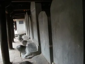 Bức ảnh chụp một dãy các bia đá lớn đặt trên lưng những con rùa đá, được xếp thẳng hàng trong một hành lang có mái che của một công trình kiến trúc cổ. Đây là hình ảnh đặc trưng của khu di tích Văn Miếu - Quốc Tử Giám tại Hà Nội, nơi có các Bia Tiến sĩ. Các bia đá được làm bằng đá, có hình chữ nhật với phần đỉnh được bo tròn. Trên bề mặt bia có khắc nhiều chữ Hán hoặc chữ Nôm, tuy nhiên không thể đọc rõ nội dung chi tiết. Mỗi bia đá được đặt trên lưng một con rùa đá được chạm khắc. Hành lang có các cột gỗ tròn màu nâu sẫm chống đỡ phần mái và nền được lát bằng gạch cũ. Ở phía xa, có thể thấy một bức tường trắng với một tấm bảng hình chữ nhật màu sẫm, nhưng không thể nhận dạng được nội dung trên đó. Không có hoạt động nào rõ rệt đang diễn ra, khung cảnh thể hiện sự tĩnh lặng, cổ kính và trang nghiêm của một di tích lịch sử.。（日本語訳）。これはハノイの文廟・國子監遺跡の特徴的な光景で、進士題名碑がある場所です。石碑は石でできており、上部が丸みを帯びた長方形をしています。それぞれの石碑は、彫刻が施された石亀の背中に乗せられています。回廊には屋根を支える濃い茶色の丸い木製の柱があり、床は古いレンガで敷き詰められています。遠くには、白い壁に濃い色の長方形の額が見えますが、その内容は識別できません。目立った活動は行われておらず、その光景は史跡の静けさ、古風さ、そして荘厳さを示しています。