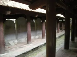 Bức ảnh chụp một hành lang có mái che trong một khu di tích lịch sử ở Hà Nội, rất có thể là khu nhà bia tại Văn Miếu - Quốc Tử Giám. Dọc theo hành lang là một dãy các bia đá lớn, mỗi bia được đặt trang trọng trên lưng một con rùa đá. Các bia và rùa này được xếp thành hàng dài. Kết cấu của hành lang bao gồm nhiều cột gỗ tròn, sơn màu nâu sẫm, chống đỡ phần mái lợp ngói. Nền nhà được lát bằng gạch vuông màu nâu đỏ. Trên bề mặt các bia đá có khắc chữ, nhưng do khoảng cách và góc chụp nên không thể đọc rõ nội dung. Khung cảnh tĩnh lặng, không có hoạt động của con người, mang đậm nét cổ kính và trang nghiêm.。（日本語訳）。写真はハノイのある史跡内にある屋根付きの廊下を写したもので、文廟・國子監の石碑のある区域である可能性が非常に高い。これらの石碑と亀は長い列に並べられている。廊下の構造は、瓦葺きの屋根部分を支える、暗褐色に塗られた多くの丸い木柱から成る。床は赤褐色の四角いレンガで敷き詰められている。石碑の表面には文字が刻まれているが、距離と撮影角度のため、内容をはっきりと読み取ることはできない。人の活動がない静かな光景は、古風で荘厳な趣を色濃く帯びている。