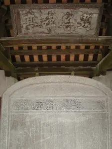 Bức ảnh chụp một tấm bia đá cổ đặt bên trong một công trình kiến trúc truyền thống, có khả năng là Văn Miếu - Quốc Tử Giám tại Hà Nội. Tấm bia đá lớn, có đỉnh hình vòm, trên bề mặt khắc dày đặc chữ Hán. Phần đầu bia có một hàng chữ Hán khổ lớn theo lối chữ triện làm tiêu đề, nhưng các ký tự đã mòn và khó nhận dạng. Phần thân bia khắc chi chít chữ nhỏ, có thể là nội dung ghi lại danh sách các tiến sĩ đỗ đạt trong các kỳ thi xưa, tuy nhiên rất khó đọc chi tiết. Phía trên tấm bia là một bức hoành phi bằng gỗ, sơn đã phai màu theo thời gian. Trên hoành phi có ba chữ Hán lớn, có thể đọc là "萬世師表" (Vạn Thế Sư Biểu), có nghĩa là "Người thầy của muôn đời". Toàn bộ tấm bia và hoành phi được đặt dưới một mái hiên có kết cấu bằng gỗ với các thanh xà và mái ngói đất nung. Khung cảnh tĩnh lặng, trang nghiêm, thể hiện sự trưng bày và bảo tồn di sản văn hóa, lịch sử.。（日本語訳）。写真は、伝統的な建築物の中に置かれた古い石碑を写したもので、ハノイの文廟・國子監である可能性が高い。大きな石碑は、上部がアーチ状になっており、表面には漢字がびっしりと刻まれている。碑の上部には、篆書体で書かれた大きな漢字が一列、題名として記されているが、文字は摩耗しており識別が困難である。碑の本体には小さな文字がびっしりと刻まれており、昔の科挙試験に合格した進士（博士）のリストを記録した内容である可能性があるが、詳細を読み取るのは非常に困難である。石碑の上には木製の扁額があり、その塗装は時間とともに色褪せている。扁額には「萬世師表」（Vạn Thế Sư Biểu）と読める大きな漢字が三文字あり、「万世の師表」という意味である。静かで荘厳な光景は、文化・歴史遺産の展示と保存を示している。