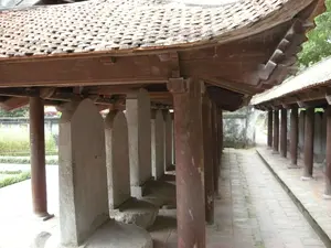 Bức ảnh chụp một hành lang có mái che bên trong một khu di tích lịch sử, có khả năng là Văn Miếu - Quốc Tử Giám ở Hà Nội. Cấu trúc này có mái ngói cong kiểu truyền thống của Việt Nam và được chống đỡ bởi các cột gỗ tròn màu nâu. Dọc theo hành lang là một dãy bia đá, mỗi bia được đặt trên lưng một con rùa đá. Đây là những tấm bia tiến sĩ, một đặc trưng của Văn Miếu. Chữ khắc trên bia không thể đọc được từ góc chụp này. Lối đi được lát gạch. Ở phía sau, có thể thấy một hành lang khác và một bức tường cũ. Không có người trong ảnh, khung cảnh rất tĩnh lặng và trang nghiêm.。（日本語訳）。この写真は、ハノイの文廟・國子監と思われる史跡内にある、屋根付きの廊下を写したものです。この建造物は、ベトナムの伝統的な様式の湾曲した瓦屋根を持ち、茶色の丸い木製の柱で支えられています。廊下に沿って石碑の列があり、それぞれの石碑は石亀の背中に乗せられています。これらは進士碑であり、文廟の特徴です。この撮影角度からは、石碑に刻まれた文字を読むことはできません。通路はタイルで舗装されています。奥には、別の廊下と古い壁が見えます。写真には人がおらず、景色は非常に静かで荘厳です。