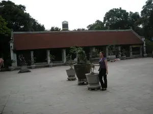 Bối cảnh là một khoảng sân rộng được lát gạch đá của một công trình kiến trúc cổ, có thể là một ngôi chùa, đình làng hoặc một di tích lịch sử. Phía sau là một dãy nhà dài, một tầng với mái ngói đỏ và hàng cột trụ màu trắng, mang kiến trúc truyền thống của Việt Nam. Một người phụ nữ, có vẻ là du khách, đang đứng trong sân bên cạnh những chậu cây cảnh lớn. Cô mặc áo không tay hoa văn màu tím và quần dài màu đen. Trong sân có đặt nhiều chậu cây cảnh được tạo dáng (bonsai). Về phía bên trái của bức ảnh, có một bệ đá với một tượng linh vật (giống sư tử đá hoặc Nghê) và một tấm bia đá có khắc chữ nhưng không thể đọc rõ. Xung quanh khu di tích có nhiều cây cối xanh tươi.。（日本語訳）。背景は、古い建築物の石畳の広い中庭で、それは寺院、村の集会所、または歴史的遺跡である可能性があります。後方には、赤い瓦屋根と白い柱列を持つ平屋の長い建物があり、ベトナムの伝統的な建築様式を呈しています。一人の女性が、観光客のようですが、大きな鉢植えの隣の中庭に立っています。彼女は紫色の柄のノースリーブのトップスと黒い長ズボンを着用しています。中庭には、形を整えられた多くの鉢植え（盆栽）が置かれています。写真の左側には、霊獣の像（石獅子またはゲに似ている）が乗った石の台座と、文字が刻まれているがはっきりと読み取れない石碑があります。史跡の周りには、青々とした木々がたくさんあります。