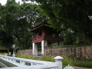 Đây là hình ảnh chụp Khuê Văn Các, một công trình biểu tượng nằm trong khu di tích Văn Miếu - Quốc Tử Giám tại Hà Nội. Khuê Văn Các là một lầu vuông hai tầng, tám mái, được xây dựng trên bốn trụ đá vững chãi. Tầng gác trên bằng gỗ, sơn màu đỏ son, có các cửa sổ hình tròn và lan can chạm khắc tinh xảo. Mái lợp ngói ống với các đầu đao cong vút. Bên dưới là một bức tường gạch cổ, rêu phong. Phía trước là một hồ nước, có thể là Hồ Văn, được bao quanh bởi lan can bằng đá màu trắng. Dọc theo bờ hồ và chân tường có nhiều cây hoa dại màu vàng và cam đang nở rộ. Khung cảnh được bao bọc bởi những cây cổ thụ lớn, tán lá xum xuê. Có một người mặc trang phục màu trắng đang đi bộ trên con đường ven hồ. Không có chữ viết nào có thể đọc được một cách rõ ràng trên công trình.。（日本語訳）。これはハノイの文廟・國子監の遺跡内にある象徴的な建築物、奎文閣を撮影した写真です。奎文閣は、頑丈な4本の石柱の上に建てられた、四角い二層八棟の楼閣です。上層階は木造で朱色に塗られ、円形の窓と精巧な彫刻が施された手すりがあります。屋根は筒瓦で葺かれ、軒先は反り上がっています。下には苔むした古いレンガの壁があります。前方には、おそらく文湖（ホーヴァン）と思われる池があり、白い石の手すりで囲まれています。池のほとりや壁の根元に沿って、黄色やオレンジ色の野生の花がたくさん咲き誇っています。周囲は、葉が青々と茂った大きな古木に囲まれています。白い服を着た人が一人、湖畔の道を歩いています。建築物の上には、はっきりと読み取れる文字はありません。