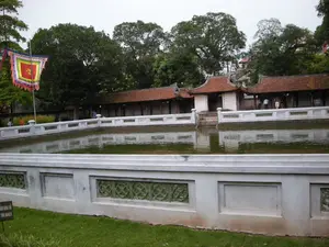 Bối cảnh là một khu di tích lịch sử, có thể là một ngôi đền hoặc văn miếu ở Hà Nội. Ở trung tâm bức ảnh là một hồ nước hình chữ nhật, mặt nước tĩnh lặng phản chiếu cảnh vật xung quanh. Bao quanh hồ là một lan can bằng đá hoặc xi măng màu trắng, được trang trí bằng các ô hoa văn màu xanh lục. Phía sau hồ là một dãy nhà hành lang mang kiến trúc cổ của Việt Nam với mái ngói đỏ và các cột gỗ. Có một vài người, dường như là khách tham quan, đang đi dạo trong hành lang. Phía sau các công trình kiến trúc là những hàng cây cổ thụ xanh tốt. Ở phía bên trái, một lá cờ hội truyền thống hình vuông, nhiều màu sắc với ngôi sao vàng ở trung tâm được treo trên một cây cột cao. Ở tiền cảnh, trên bãi cỏ, có một tấm biển nhỏ màu đen ghi dòng chữ: "XIN ĐỪNG LÊN CỎ" và bên dưới là "KEEP OFF THE GRASS".。（日本語訳）。背景は歴史的遺跡で、ハノイの寺院か文廟（ぶんびょう）かもしれません。写真の中央には長方形の池があり、静かな水面が周囲の景色を映し出しています。池の周りには石またはセメントでできた白い欄干があり、緑色の模様のパネルで装飾されています。池の後ろには、赤い瓦屋根と木製の柱を持つ、ベトナムの古い建築様式の回廊が並んでいます。回廊の中を、観光客と思われる数人の人々が散策しています。建築物の後ろには、青々と茂る古い木々が並んでいます。左側には、高い竿に、中央に黄色い星が描かれた色とりどりの四角い伝統的な祭りの旗が掲げられています。前景の芝生の上には、「XIN ĐỪNG LÊN CỎ」という文字と、その下に「KEEP OFF THE GRASS」と書かれた小さな黒い看板があります。