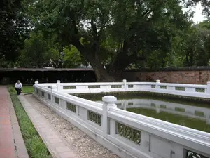 Bức ảnh chụp một khung cảnh thanh bình bên trong một khu di tích lịch sử, có thể là một ngôi đền ở Hà Nội. Ở trung tâm là một hồ nước nhỏ hình chữ nhật, được bao quanh bởi lan can xi măng sơn trắng với các họa tiết trang trí. Nước trong hồ có màu xanh lục. Dọc theo hồ là một lối đi lát gạch và một lối đi rải sỏi, ngăn cách bởi một dải cỏ. Ở phía bên trái, một người đàn ông mặc áo sơ mi trắng đang ngồi xổm trên bãi cỏ. Phía sau là một cây cổ thụ lớn với tán lá xum xuê. Trong hậu cảnh, có thể thấy một bức tường gạch cũ và một khu vực có mái che, trông giống như một bãi đỗ xe với nhiều xe máy đang đậu.。（日本語訳）。写真は、歴史的遺跡の内部の穏やかな風景を写しており、ハノイのある寺院かもしれない。中央には長方形の小さな池があり、装飾的な模様が施された白塗りのセメント製の手すりで囲まれている。池の水は緑色である。池に沿って、レンガ敷きの通路と砂利の通路があり、それらは芝生の帯で隔てられている。左側では、白いシャツを着た男性が芝生の上でしゃがんでいる。後方には、葉が青々と茂った大きな古木がある。