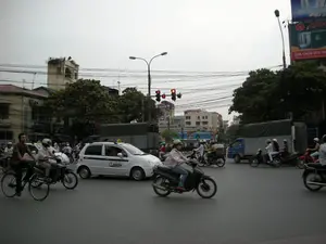 Đây là một cảnh giao thông đường phố tại một ngã tư đông đúc ở Hà Nội. Bầu trời u ám. Rất nhiều người đang tham gia giao thông với các phương tiện đa dạng, chủ yếu là xe máy. Hầu hết người đi xe máy đều đội mũ bảo hiểm, và một số người đeo khẩu trang. Ở giữa bức ảnh, một chiếc taxi màu trắng đang di chuyển. Ngoài ra còn có một chiếc xe đạp, một xe tải màu xanh và một xe tải quân sự phủ bạt. Đèn tín hiệu giao thông đang ở trạng thái màu đỏ. Hệ thống dây điện chằng chịt vắt ngang trên đường phố. Hai bên đường là các tòa nhà và cây xanh. Ở phía bên phải có một tấm biển quảng cáo lớn với dòng chữ 'LỰA CHỌN DUY NHẤT' và tên thương hiệu 'APPO...' bị che một phần. Ở phía bên trái, có một biển hiệu khác ghi 'TÂN &...' nhưng không thể đọc được toàn bộ.。（日本語訳）。これはハノイの混雑した交差点における路上交通の光景です。空は曇っています。多くの人々が、主にバイクといった多様な乗り物で交通に参加しています。バイクに乗る人のほとんどはヘルメットを着用しており、何人かはマスクをしています。写真の中央では、白いタクシーが移動しています。その他にも、自転車一台、青いトラック一台、そして幌で覆われた軍用トラック一台があります。交通信号は赤です。複雑に絡み合った電線が通りを横切っています。道路の両側には建物と緑の木々があります。右側には、「LỰA CHỌN DUY NHẤT」という文字と、一部が隠れているブランド名「APPO...」が書かれた大きな広告看板があります。左側には、「TÂN &...」と書かれた別の看板がありますが、全部を読むことはできません。