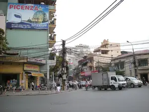 Bối cảnh là một ngã tư đường phố tại Hà Nội. Nổi bật trên một tòa nhà cao tầng màu xanh lá cây là tấm biển quảng cáo lớn của thương hiệu PICENZA với nội dung: "PICENZA", "Bình nước nóng tráng men - TITANIUM", "TẬP ĐOÀN PICENZA - VIỆT NAM", và trang web "WWW.PICENZA.COM.VN". Phía dưới, ở tầng trệt, là một cửa hàng có biển hiệu màu vàng ghi "PHOTO COPY" và dòng chữ nhỏ hơn "ĐÓNG GÁY XOẮN - INA CHỮ VÀNG". Một biển hiệu khác cũng ghi "PHOTOCOPY". Khung cảnh đường phố tấp nập với nhiều phương tiện giao thông đang di chuyển, bao gồm nhiều xe máy, ô tô và một chiếc xe tải nhỏ màu trắng. Một đặc điểm nổi bật của cảnh quan đô thị là hệ thống dây điện và dây cáp chằng chịt, rối bù trên các cột điện. Các tòa nhà là kiểu nhà ống điển hình, san sát nhau. Một lá cờ Việt Nam được treo trên một cột. Một vài người đang ngồi trên vỉa hè trước các cửa hàng.。（日本語訳）。背景はハノイのある街角の交差点です。緑色の高層ビルには、PICENZAブランドの大きな広告看板が目立っており、その内容は「PICENZA」、「ホーロー製給湯器 - TITANIUM」、「PICENZAグループ - ベトナム」、そしてウェブサイト「WWW.PICENZA.COM.VN」です。その下、1階には「PHOTO COPY」と書かれた黄色い看板の店があり、より小さな文字で「スパイラル製本 - 金文字印刷」と書かれています。別の看板にも「PHOTOCOPY」と書かれています。街の風景は、多くのバイク、自動車、そして一台の白い小型トラックを含む、移動中の多くの乗り物で賑わっています。都市景観の際立った特徴は、電柱の上で複雑に絡み合った電線とケーブル網です。建物は典型的なチューブハウスで、隣り合って密集しています。一本の柱にベトナム国旗が掲げられています。数人が店の前の歩道に座っています。