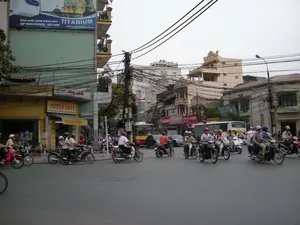 Đây là hình ảnh một góc phố hoặc ngã tư đông đúc tại một thành phố ở Việt Nam, có thể là Hà Nội. Hoạt động chính là giao thông đường bộ, với rất nhiều người đang điều khiển xe máy trên đường. Hầu hết người đi xe máy đều đội mũ bảo hiểm. Dòng xe dường như đang dừng lại hoặc di chuyển chậm. Ngoài xe máy, còn có một chiếc xe buýt màu trắng và một vài chiếc ô tô cũng tham gia giao thông. Các tòa nhà là kiểu nhà ống nhiều tầng, san sát nhau. Một đặc điểm nổi bật là hệ thống dây điện và cáp viễn thông chằng chịt trên các cột điện. Có một số biển hiệu có thể đọc được: trên một tòa nhà cao tầng có tấm biển quảng cáo lớn màu xanh ghi 'Bình nước nóng tráng men - TITANIUM', 'TẬP ĐOÀN PICENZA - VIỆT NAM', và website 'WWW.PICENZA.COM.VN'. Ở tầng trệt của một tòa nhà khác có một cửa hàng với biển hiệu màu vàng ghi 'PHOTO COPY'.。（日本語訳）。これはベトナムのある都市、おそらくハノイの、賑やかな街角または交差点の画像です。主な活動は道路交通で、非常に多くの人々が道路でバイクを運転しています。バイクに乗る人のほとんどはヘルメットを着用しています。車両の流れは停止しているか、ゆっくりと動いているようです。バイクの他に、白いバス1台と数台の自動車も交通に参加しています。建物は多層階のチューブハウス（間口が狭く奥行きが深い家）で、互いに密集しています。際立った特徴は、電柱に張り巡らされた電線と通信ケーブルのシステムです。いくつか読み取れる看板があります。高層ビルには、「Bình nước nóng tráng men - TITANIUM」（チタンエナメル温水器）、「TẬP ĐOÀN PICENZA - VIỆT NAM」（ピチェンツァグループ - ベトナム）、そしてウェブサイト「WWW.PICENZA.COM.VN」と書かれた大きな青い広告看板があります。別の建物の1階には、「PHOTO COPY」と書かれた黄色い看板のある店があります。