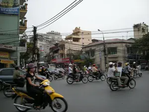 Các dòng chữ có thể đọc được trên các biển hiệu bao gồm: 'TITANIUM' và 'WEBSITE: WWW.PICENZA.COM.VN' trên một tòa nhà màu xanh lam ở bên trái; 'TO COPY' và 'PHOTOCOPY' trên các biển hiệu của một cửa hàng. Có nhiều biển hiệu khác ở phía xa nhưng không thể đọc rõ chữ.。Đây là một cảnh đường phố đông đúc tại một thành phố, có khả năng là Hà Nội. Bối cảnh là một ngã tư hoặc một con đường lớn với mật độ giao thông cao, đặc trưng bởi dòng xe máy dày đặc. Hoạt động chính là mọi người đang tham gia giao thông. Rất nhiều người đang điều khiển xe máy (xe số, xe ga), tất cả đều đội mũ bảo hiểm. Một số người, như người phụ nữ lái chiếc xe máy màu vàng ở phía trước, đeo khẩu trang. Ngoài xe máy, còn có ô tô và một chiếc xe buýt màu trắng ở phía sau. Các tòa nhà là kiểu nhà ống cao tầng đặc trưng của đô thị Việt Nam, được xây san sát nhau. Một đặc điểm nổi bật của khung cảnh là hệ thống dây điện và cáp viễn thông chằng chịt, treo trên các cột điện. Bầu trời có vẻ u ám.。（日本語訳）。看板で読み取れる文字には、左側の青い建物にある「TITANIUM」と「WEBSITE: WWW.PICENZA.COM.VN」、ある店の看板にある「TO COPY」と「PHOTOCOPY」が含まれます。遠くには他にも多くの看板がありますが、文字ははっきりと読み取れません。これはある都市の混雑した街頭の風景で、ハノイである可能性があります。背景は交差点か、または交通量の多い大通りで、びっしりと並んだバイクの流れが特徴です。主な活動は、人々が交通に参加していることです。非常に多くの人々がバイク（マニュアル車、スクーター）を運転しており、全員がヘルメットを着用しています。手前の黄色いバイクを運転している女性のように、一部の人々はマスクを着用しています。バイクの他に、自動車や後方には白いバスも見られます。建物はベトナムの都市に特有の、隣接して建てられた高層のチューブハウス（縦長の家）です。この風景の際立った特徴は、電柱にぶら下がっている、複雑に絡み合った電線と通信ケーブルのシステムです。空は曇っているようです。