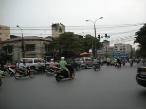 Đây là một cảnh giao thông đường phố tại một khu vực đô thị ở Hà Nội. Con đường đông đúc với nhiều phương tiện đang di chuyển, chủ yếu là xe máy và một số ô tô. Đèn tín hiệu giao thông đang bật màu xanh, cho thấy các phương tiện đang được phép đi. Hầu hết những người điều khiển xe máy đều đội mũ bảo hiểm. Ở phía xa, có thể thấy một người đi xe đạp. Hai bên đường là các tòa nhà nhiều tầng, có cả nhà ở và cửa hàng. Một đặc điểm nổi bật của khung cảnh là hệ thống dây điện và dây cáp chằng chịt ở phía trên. Bầu trời u ám. Có một số biển hiệu trên các tòa nhà nhưng chữ viết không thể đọc rõ. Một biển hiệu màu xanh dường như có chữ 'TÂN &...'. Phía trước một chiếc ô tô có thể thấy một phần chữ 'NIS...'.。（日本語訳）。これはハノイの都市部における路上交通の一場面です。道路は多くの乗り物で混雑しており、主にバイクと数台の自動車が移動しています。交通信号は青色で、車両の通行が許可されていることを示しています。バイクを運転している人のほとんどはヘルメットを着用しています。遠くには、自転車に乗っている人が一人見えます。道路の両側には、住宅と店舗の両方を含む多層階の建物が並んでいます。この光景の際立った特徴は、上部に複雑に張り巡らされた電線とケーブルです。空は曇っています。建物にはいくつかの看板がありますが、文字ははっきりと読み取れません。青い看板には「TÂN &...」という文字があるようです。一台の自動車の前方に、「NIS...」という文字の一部が見えます。