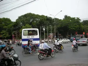 Bức ảnh chụp cảnh giao thông tại một ngã tư đường phố đông đúc ở một thành phố, có thể là Hà Nội. Dòng phương tiện rất đa dạng, bao gồm rất nhiều xe máy, một chiếc xe buýt nhỏ màu trắng và xanh dương, một vài chiếc ô tô con và cả xe đạp. Mọi người đang điều khiển phương tiện của mình để di chuyển qua ngã tư. Hầu hết người đi xe máy đều đội mũ bảo hiểm. Ở bên đường có thể thấy các biển báo giao thông, bao gồm biển báo cấm đỗ xe và biển báo cấm đi ngược chiều. Phía trên là hệ thống dây điện chằng chịt. Phông nền là những hàng cây xanh lớn, tạo nên một khung cảnh đô thị đặc trưng của Việt Nam.。（日本語訳）。ある都市、おそらくハノイの、混雑した街路の交差点での交通風景を撮った写真です。車両の流れは非常に多様で、非常に多くのバイク、一台の白と青色の小型バス、数台の乗用車、そして自転車も含まれています。人々は交差点を渡るために自分の乗り物を運転しています。バイクに乗る人のほとんどはヘルメットをかぶっています。道端には、駐車禁止の標識や逆走禁止の標識を含む交通標識が見えます。上の方には、複雑に絡み合った電線があります。背景には大きな緑の並木があり、ベトナム特有の都市の光景を作り出しています。