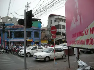 Bối cảnh là một ngã tư đường phố đông đúc tại Hà Nội. Nhiều phương tiện giao thông, chủ yếu là xe máy và một vài chiếc ô tô (bao gồm cả taxi), đang dừng chờ đèn tín hiệu. Hầu hết những người đi xe máy đều đội mũ bảo hiểm. Phía bên phải có một tấm áp phích lớn màu hồng với dòng chữ 'CHÀO MỪNG KỶ NIỆM... VÀ QUỐC KHÁNH NƯỚC...', cho thấy không khí chào mừng một ngày lễ lớn của quốc gia. Ở phía đối diện, có một tòa nhà màu xanh lam là cửa hàng nội thất với các biển hiệu: 'Hapro NỘI THẤT VĂN PHÒNG - GIA ĐÌNH - TRƯỜNG HỌC' và băng rôn 'KHAI TRƯƠNG HỆ THỐNG 16 CỬA HÀNG CHUYÊN...'. Gần đó có một biển báo giao thông 'CẤM ĐI NGƯỢC CHIỀU' và biển chỉ dẫn 'PHỐ BIA', 'MÓN XÀO'. Khung cảnh đặc trưng của đô thị Việt Nam với hệ thống dây điện chằng chịt trên cao và các tòa nhà san sát. Trên cột đèn giao thông có gắn một vật trang trí hình bông tuyết.。（日本語訳）。背景はハノイの混雑した交差点です。多くの交通手段、主にバイクと数台の自動車（タクシーを含む）が信号待ちをしています。バイクに乗っている人のほとんどはヘルメットを着用しています。右側には「...記念日と国家の建国記念日をお祝いします」という文字が書かれた大きなピンク色のポスターがあり、国の大きな祝日を祝う雰囲気を示しています。反対側には青い建物があり、それは「Hapro オフィス・家庭・学校用家具」という看板と「専門16店舗システム開店...」という横断幕がある家具店です。その近くには、「逆走禁止」という交通標識と、「ビアストリート」、「炒め物」という案内看板があります。頭上に張り巡らされた電線網と密集した建物が特徴的なベトナムの都市の光景です。信号機の柱には、雪の結晶の形をした装飾品が取り付けられています。