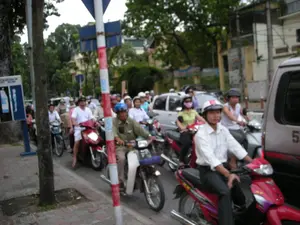 Bức ảnh chụp lại một cảnh tượng giao thông đông đúc trên một con đường ở thành phố, có khả năng là Hà Nội. Một hàng dài xe máy đang nối đuôi nhau, có vẻ như đang dừng lại do tắc đường. Người điều khiển xe máy bao gồm cả nam và nữ, với nhiều độ tuổi khác nhau, và hầu hết mọi người đều tuân thủ quy định đội mũ bảo hiểm. Một số người đeo khẩu trang để tránh khói bụi. Ở phía trước, một người đàn ông mặc áo sơ mi trắng đang ngồi trên chiếc xe máy màu đỏ. Bên cạnh anh là những người đi xe máy khác, và ở bên phải là một chiếc xe ô tô tải van màu bạc. Dọc vỉa hè, có một cây cột được sơn sọc đỏ và trắng, là một loại cọc tiêu giao thông. Gần đó có một biển báo công cộng màu xanh dương, có thể là biển báo cho bốt điện thoại công cộng, nhưng chữ viết trên đó khá mờ và khó đọc. Bối cảnh phía sau là những hàng cây xanh và các tòa nhà đặc trưng của một khu đô thị.。（日本語訳）。写真は、都市の道路での混雑した交通風景を捉えており、ハノイである可能性が高い。長いバイクの列が連なっており、交通渋滞で停止しているようだ。バイクの運転手には男性も女性も含まれ、年齢層も様々で、ほとんどの人がヘルメット着用規定を遵守している。排気ガスや埃を避けるためにマスクを着用している人もいる。前方では、白いシャツを着た男性が赤いバイクに乗っている。彼の隣には他のバイク運転手がおり、右側には銀色のバンがいる。歩道沿いには、赤と白の縞模様に塗られた柱があり、これは交通用の標柱の一種である。その近くには青い公共の標識があり、公衆電話ボックスの標識かもしれないが、そこに書かれている文字はかなりぼやけていて読みにくい。
