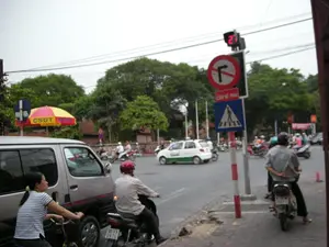 Bối cảnh là một ngã tư đường phố tại Hà Nội. Dòng người và các phương tiện giao thông đang dừng chờ đèn đỏ. Có nhiều người đi xe máy, một người phụ nữ đi xe đạp và một chiếc ô tô tải nhỏ màu bạc đang ở gần lề đường. Mọi người đi xe máy đều đội mũ bảo hiểm. Trên cột tín hiệu giao thông, có một đồng hồ đếm ngược đang hiển thị số "21". Ngay dưới đó là một biển báo hình tròn viền đỏ cấm rẽ phải, và một biển phụ hình chữ nhật ghi rõ "CẤM RẼ PHẢI". Dưới cùng là biển báo màu xanh chỉ lối cho người đi bộ qua đường. Ở phía bên kia đường, có một chiếc ô lớn màu vàng và đỏ với dòng chữ "CSGT HÀ NỘI", cho thấy đây có thể là một chốt của Cảnh sát Giao thông. Phía sau là khung cảnh đường phố với nhiều cây xanh và các phương tiện khác đang lưu thông, bao gồm một chiếc taxi.。（日本語訳）。背景はハノイのある街の交差点です。人々と車両の流れは赤信号で止まり、待っています。たくさんのバイクに乗った人々、自転車に乗った一人の女性、そして銀色の小型トラック一台が路肩近くにいます。バイクに乗っている人は皆ヘルメットを着用しています。信号機の柱には、「21」という数字を表示しているカウントダウン時計があります。そのすぐ下には、右折を禁止する赤枠の円形標識と、「CẤM RẼ PHẢI」（右折禁止）と明記された長方形の補助標識があります。一番下には、歩行者横断用の通路を示す青い標識があります。道路の向こう側には、「CSGT HÀ NỘI」という文字が入った黄色と赤の大きな傘があり、これが交通警察の詰め所である可能性を示しています。後方には、多くの緑の木々や、タクシー一台を含む他の車両が通行している街の風景が広がっています。