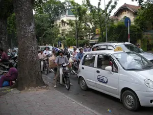 Trên xe taxi màu trắng có ghi dòng chữ 'VIET THANH TAXI' và số điện thoại '8.39.39.39'. Trên nóc xe có biển hiệu 'TAXI'. Ở phía sau, trên một tòa nhà có biển hiệu màu vàng ghi chữ 'HÒA GIẶT C...', có thể là một cửa hàng giặt là.。Bối cảnh là một con đường trong thành phố với nhiều cây cổ thụ lớn ở hai bên vỉa hè. Dòng phương tiện giao thông đang đông đúc, bao gồm rất nhiều xe máy và ô tô, dường như đang dừng lại hoặc di chuyển rất chậm, cho thấy khả năng đang xảy ra ùn tắc giao thông. Hầu hết những người đi xe máy đều đội mũ bảo hiểm. Một chiếc xe taxi màu trắng nằm ở phía trước, bên cạnh là dòng xe máy. Một người đàn ông đi xe máy đang chở một túi giấy lớn. Ở bên lề đường, dưới gốc cây, một người phụ nữ mặc đồ màu tím đang ngồi trên một chiếc ghế đẩu nhỏ. Phía sau là các tòa nhà nhiều tầng và cây cối um tùm. Có một biển báo giao thông hình tròn màu xanh lam.。（日本語訳）。白いタクシーには「VIET THANH TAXI」という文字と電話番号「8.39.39.39」が書かれています。車の屋根には「TAXI」という表示があります。後方には、ある建物に「HÒA GIẶT C...」と書かれた黄色い看板があり、クリーニング店かもしれません。背景は都市の道路で、歩道の両側には多くの大きな古木があります。交通量は多く、多くのバイクや自動車が含まれており、停止しているか、非常にゆっくりと動いているように見え、交通渋滞が発生している可能性を示しています。バイクに乗っている人のほとんどはヘルメットを着用しています。前方には白いタクシーが1台あり、その隣にはバイクの列があります。バイクに乗った男性が大きな紙袋を運んでいます。道端の木陰では、紫色の服を着た女性が小さなスツールに座っています。後方には高層ビルや生い茂る木々があります。青い円形の交通標識が1つあります。