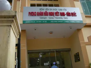 Bức ảnh chụp lối vào của một cơ sở y tế, cụ thể là Phòng khám Hữu nghị Việt Nam - Hàn Quốc, thuộc Bệnh viện Đa khoa Xanh Pôn tại Hà Nội.。**Thông tin chữ viết được ghi lại:**。Trên biển hiệu lớn có các dòng chữ sau:。- Dòng trên cùng: BỆNH VIỆN ĐA KHOA XANH PÔN。- Dòng chữ lớn màu đỏ: PHÒNG KHÁM HỮU NGHỊ VIỆT NAM - HÀN QUỐC。- Dòng chữ tiếng Anh: Vietnam - Korea Friendship Clinic。- Trên dải màu xanh lá cây ở dưới cùng:。- Địa chỉ: 12 CHU VĂN AN - BA ĐÌNH - HÀ NỘI。- Số điện thoại: ĐIỆN THOẠI: 8 437231 - 7.346835 - 7.346836 - 7.346837。- Trên cửa kính có dán giấy ghi chữ "Kéo ra".。**Miêu tả cảnh vật và hoạt động:**。- **Loại địa điểm:** Lối vào một phòng khám y tế nằm trong một tòa nhà được sơn màu vàng. Đây là một phần của một bệnh viện lớn hơn.。- **Hoạt động cụ thể:** Một người đàn ông mặc áo xanh và đội mũ đang đứng ở cửa ra vào bằng kính, dường như đang mở cửa để đi vào hoặc đi ra khỏi phòng khám.。- **Vật thể trong ảnh:** Một biển hiệu lớn của phòng khám, một tòa nhà có kiến trúc hiện đại, cửa ra vào bằng kính, một cột cổng bằng bê tông ở bên trái, và một phần của cổng sắt màu đen. Dưới mái che của lối vào có lắp đặt các bóng đèn trần.。（日本語訳）。写真は、ハノイにあるサンポール総合病院に属する医療施設、具体的にはベトナム-韓国友好診療所の入口を撮影したものです。**記録されている文字情報：**。大きな看板には以下の文言があります：。- 最上段の行：BỆNH VIỆN ĐA KHOA XANH PÔN。- 赤色の大きな文字の行：PHÒNG KHÁM HỮU NGHỊ VIỆT NAM - HÀN QUỐC。- 英語の行：Vietnam - Korea Friendship Clinic。- 一番下の緑色の帯の上：。- 住所：12 CHU VĂN AN - BA ĐÌNH - HÀ NỘI。- 電話番号：ĐIỆN THOẠI: 8 437231 - 7.346835 - 7.346836 - 7.346837。- ガラスのドアには「Kéo ra」（引く）と書かれた紙が貼られています。**風景と活動の描写：**。- **場所の種類：** 黄色に塗られた建物内にある医療診療所の入口。これは、より大きな病院の一部です。- **具体的な活動：** 青いシャツを着て帽子をかぶった男性がガラスの出入口に立っており、診療所に入るか出るためにドアを開けているように見えます。- **写真の中の物体：** 診療所の大きな看板、近代的な建築様式の建物、ガラスの出入口、左側にあるコンクリート製の門柱、そして黒い鉄製の門の一部。入口の庇の下には天井灯が設置されています。