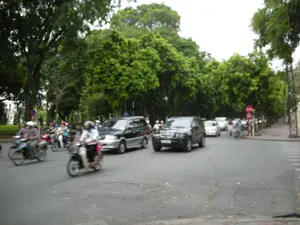 Bức ảnh chụp một cảnh giao thông trên một con đường ở Hà Nội. Con đường được trải nhựa, hai bên là hàng cây xanh lớn rợp bóng mát. Nhiều phương tiện đang lưu thông trên đường, bao gồm rất nhiều xe máy và một vài chiếc ô tô. Những người điều khiển xe máy đều đội mũ bảo hiểm. Ở phía bên trái có một biển báo giao thông hình tròn màu đỏ là biển "Cấm đi ngược chiều". Phía xa bên phải cũng có một biển báo giao thông hình tròn màu đỏ khác nhưng không thể nhìn rõ ký hiệu. Trên mặt đường bên phải có vạch kẻ đường dành cho người đi bộ.。（日本語訳）。写真はハノイのある道路の交通風景を撮影したものです。道路は舗装されており、両側には大きな緑の木々が並び、涼しい木陰を作っています。道路上では多くの乗り物が通行しており、その中にはたくさんのバイクと数台の自動車が含まれています。バイクを運転している人々は皆ヘルメットを着用しています。左側には、「一方通行（進入禁止）」を示す赤色の円形交通標識があります。右側の遠くにも別の赤色の円形交通標識がありますが、記号ははっきりと見えません。右側の路面には、歩行者用の横断歩道があります。