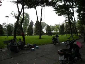 Bối cảnh là một công viên công cộng ở Hà Nội, với nhiều cây xanh lớn, bãi cỏ và lối đi bộ. Ở phía xa, có một tượng đài lớn của một người đàn ông đứng trên bệ. Ở phía trước, một nhóm ba người đàn ông đang ngồi trên những chiếc ghế nhựa thấp, tụ tập quanh một quán nước vỉa hè tạm bợ được bày ra trên mặt đất. Họ đang uống nước và trò chuyện. Đây là một hoạt động thư giãn thường thấy trong các công viên. Có một vài chiếc xe máy và một chiếc xe đạp đang đỗ gần đó. Một chiếc xe máy ở tiền cảnh có treo một chiếc túi ni lông màu hồng. Trong công viên còn có ghế đá và cột đèn đường. Không có chữ viết hay biển hiệu nào có thể đọc được.。（日本語訳）。背景はハノイのある公共公園で、大きな緑の木々、芝生、遊歩道がたくさんあります。遠くには、台座の上に立つ男性の大きな記念碑があります。手前では、3人の男性のグループが低いプラスチックの椅子に座り、地面に広げられた臨時の歩道の飲み物屋台の周りに集まっています。彼らは飲み物を飲みながらおしゃべりをしています。これは公園でよく見られるリラックスするための活動です。その近くに数台のバイクと一台の自転車が停まっています。前景にあるバイクには、ピンク色のビニール袋が掛かっています。公園の中には石のベンチや街灯もあります。読み取れる文字や看板はありません。