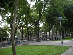 Bối cảnh là một công viên hoặc một không gian xanh ven một con đường ở thành phố. Con đường được rợp bóng bởi nhiều cây cổ thụ to lớn, tán lá xum xuê. Phía trước là một bãi cỏ xanh. Trên đường, có nhiều phương tiện giao thông đang di chuyển, bao gồm xe máy và một chiếc xe tải nhỏ màu trắng. Trên vỉa hè, một nhóm người đang tụ tập, một số ngồi trên ghế, có vẻ như đang trò chuyện hoặc nghỉ ngơi. Một vài người khác đang đi bộ. Có một cây đèn đường với thiết kế đặc trưng. Ở phía xa, có thể thấy một cổng vào với kiến trúc cổ và một tòa nhà được bao quanh bởi hàng rào. Có các biển báo nhưng chữ viết trên đó không thể đọc được.。（日本語訳）。背景は公園、あるいは都市の道沿いにある緑地です。道は、葉が生い茂る多くの大きな古木によって影ができています。手前には緑の芝生があります。道路上では、バイクや白い小型トラックなど、多くの交通車両が移動しています。歩道では、人々のグループが集まっており、何人かは椅子に座って、おしゃべりをしたり休憩したりしているようです。他の数人が歩いています。特徴的なデザインの街灯があります。遠くには、古い建築様式の門と、柵で囲まれた建物が見えます。標識がありますが、そこに書かれている文字は読めません。