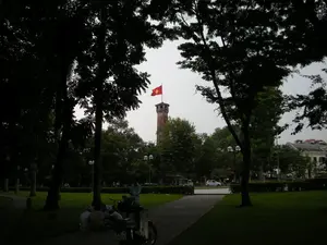 Bức ảnh chụp một khung cảnh trong công viên ở Hà Nội. Ở trung tâm phía xa là Cột cờ Hà Nội, một tòa tháp bằng gạch cổ kính, trên đỉnh đang tung bay lá cờ đỏ sao vàng của Việt Nam. Xung quanh là rất nhiều cây cổ thụ lớn với tán lá rậm rạp, tạo ra những khoảng tối và bóng mát. Ở phía trước, dưới bóng cây, một nhóm người đang ngồi nghỉ ngơi trên bãi cỏ bên cạnh một lối đi bộ. Gần đó có một chiếc xe máy đang dựng. Phía sau, qua những hàng cây, có thể thấy một con đường với vài chiếc ô tô và một số tòa nhà. Không có văn bản hay chữ viết nào có thể nhận dạng được trong ảnh.。（日本語訳）。この写真はハノイの公園の風景を撮影したものです。遠く中央にはハノイの旗竿塔（コッコー・ハノイ）があります。これは古いレンガ造りの塔で、その頂上にはベトナムの金星紅旗がはためいています。周囲には、葉が鬱蒼と茂った多くの大きな古木があり、暗い部分や木陰を作り出しています。手前では、木陰の下、遊歩道の隣の芝生の上で、数人のグループが座って休んでいます。その近くにバイクが1台停まっています。後方には、木々の向こうに、数台の車といくつかの建物がある道路が見えます。写真の中には、認識できるテキストや文字はありません。