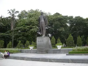 Trên bệ tượng có khắc dòng chữ: "V.I. LÊ-NIN". Bức ảnh chụp một tượng đài lớn đặt tại một công viên công cộng, có thể là Công viên Lê-nin ở Hà Nội. Bức tượng bằng đồng khắc họa hình ảnh V.I. Lê-nin đang đứng, được đặt trên một bệ đá lớn. Xung quanh tượng đài là một quảng trường được lát đá, có các bồn hoa được chăm sóc cẩn thận và nhiều cây xanh rậm rạp ở phía sau, trong đó có một cây cọ cao. Ở phía bên trái, hai người phụ nữ đang ngồi nghỉ ngơi trên bậc thềm đá, bên cạnh có một chiếc xe đạp dựng. Khung cảnh cho thấy một không gian công cộng yên tĩnh.。（日本語訳）。台座には「V.I. レーニン」という文字が刻まれている。この写真は、ハノイのレーニン公園と思われる公共の公園に設置された大きな記念碑を写している。記念碑の周りは石畳の広場になっており、手入れの行き届いた花壇や、後方には背の高いヤシの木を含む多くの緑豊かな木々がある。左側では、二人の女性が石段に座って休んでおり、その隣には自転車が一台立てかけられている。この光景は、静かな公共空間を示している。