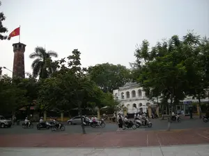 Bức ảnh chụp một cảnh đường phố ở Hà Nội. Nổi bật ở phía bên trái là Cột cờ Hà Nội, một tòa tháp lịch sử, trên đỉnh đang tung bay lá cờ đỏ sao vàng của Việt Nam. Con đường nhựa đông đúc với nhiều người đang tham gia giao thông, chủ yếu bằng xe máy, một phương tiện đi lại phổ biến. Một số người lái xe máy có đội mũ bảo hiểm. Có cả ô tô đang lưu thông trên đường. Dọc theo con đường và xung quanh Cột cờ có rất nhiều cây xanh tươi tốt. Ở phía sau, có thể thấy một tòa nhà lớn màu trắng với kiến trúc kiểu Pháp cổ, có các cửa sổ hình vòm. Vỉa hè được lát gạch đỏ. Có một số biển hiệu trên các tòa nhà nhưng không thể đọc rõ chữ.。（日本語訳）。写真はハノイのある街の風景を撮影したものです。左側で際立っているのは、歴史的な塔であるハノイの旗竿であり、その頂上にはベトナムの金星紅旗がはためいています。アスファルトの道路は多くの交通参加者で混雑しており、主に一般的な交通手段であるバイクを利用しています。一部のバイク運転手はヘルメットを着用しています。道路には自動車も走行しています。道路沿いと旗竿の周りには、青々とした緑の木々がたくさんあります。後方には、アーチ型の窓がある、古いフランス様式の建築を持つ大きな白い建物が見えます。歩道は赤いレンガで舗装されています。