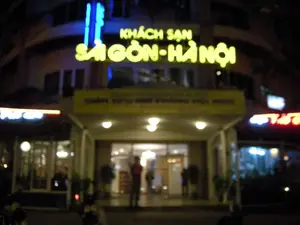 Bức ảnh chụp cảnh ban đêm ở lối vào một khách sạn. Một biển hiệu lớn có đèn màu vàng ghi rõ dòng chữ 'KHÁCH SẠN SÀI GÒN - HÀ NỘI'. Ngay phía dưới, trên mái che lối vào, có một băng rôn màu vàng nhưng chữ bị mờ, khó đọc, chỉ có thể nhận ra một vài từ như '... PHÒNG HỘI NGHỊ'. Lối vào chính được chiếu sáng bởi nhiều ngọn đèn. Bên trong cửa ra vào, có một người đàn ông mặc áo đỏ đang đứng. Phía trước tòa nhà có một vài chiếc xe máy đang đỗ. Hai bên lối vào cũng có các cửa hàng hoặc các khu vực khác của khách sạn với biển hiệu đèn neon nhưng không thể đọc rõ chữ.。（日本語訳）。あるホテルの入り口の夜景を写した写真です。黄色い電飾の大きな看板に「KHÁCH SẠN SÀI GÒN - HÀ NỘI」という文字がはっきりと書かれています。そのすぐ下、入り口の庇の上には黄色い横断幕がありますが、文字はぼやけていて読みにくく、「... PHÒNG HỘI NGHỊ」のような単語がいくつか認識できるだけです。正面玄関は多くの照明で照らされています。入り口のドアの内側には、赤いシャツを着た男性が立っています。建物の前には数台のバイクが駐車されています。