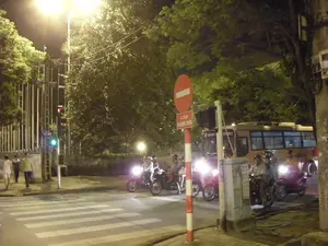 Đây là cảnh một ngã tư đường phố ở Hà Nội vào ban đêm. Nhiều người đi xe máy, tất cả đều đội mũ bảo hiểm, đang dừng lại trước vạch kẻ đường cho người đi bộ và chờ tín hiệu giao thông. Phía sau họ có một chiếc xe buýt màu vàng. Bên lề đường, một vài người đi bộ đang đi qua. Con đường được chiếu sáng bởi ánh đèn đường vàng, và hai bên đường có nhiều cây xanh. Có một biển báo giao thông cấm đi ngược chiều với dòng chữ "CẤM ĐI NGƯỢC CHIỀU". Một biển tên đường màu xanh lam có thể được nhìn thấy với dòng chữ "YẾT KIÊU". Đèn tín hiệu giao thông cho người đi bộ đang bật màu xanh.。（日本語訳）。これは夜のハノイの街の交差点の光景です。多くのバイクに乗った人々が、全員ヘルメットをかぶり、横断歩道の手前で停止し、信号を待っています。彼らの後ろには黄色いバスが1台います。道端では、数人の歩行者が渡っています。道路は黄色い街灯で照らされており、道の両側には多くの緑の木々があります。「CẤM ĐI NGƯỢC CHIỀU」という文字が書かれた、逆走を禁止する交通標識があります。「YẾT KIÊU」という文字が書かれた青い道路名の標識が見えます。歩行者用の信号が青になっています。