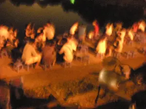 Bức ảnh chụp một cảnh tượng vào ban đêm, hình ảnh khá mờ và nhiễu. Có một nhóm đông người đang tụ tập ở một không gian ngoài trời, có thể là vỉa hè hoặc một khu vực công cộng. Mọi người đang ngồi trên những chiếc ghế nhựa thấp, một hình ảnh rất đặc trưng của các quán nước hay quán ăn vỉa hè ở Hà Nội. Họ dường như đang trò chuyện và giao lưu với nhau. Ánh sáng màu vàng ấm áp từ đèn đường hoặc đèn của quán chiếu sáng khu vực, tạo nên một không khí tụ tập về đêm. Do chất lượng ảnh thấp và bị nhòe, không thể nhận dạng được khuôn mặt, các vật thể cụ thể hay đọc được bất kỳ chữ viết nào trên biển hiệu nếu có.。（日本語訳）。夜の光景を写した写真で、画像はかなりぼやけており、ノイズが入っています。大勢の人が屋外の空間に集まっています。それは歩道か、あるいは公共の場所かもしれません。人々は低いプラスチックの椅子に座っており、これはハノイの路上にある飲み物屋や食堂の非常に特徴的な光景です。彼らはお互いに会話をしたり、交流したりしているようです。街灯や店の灯りからの暖かい黄色の光がそのエリアを照らし、夜の集まりの雰囲気を醸し出しています。写真の画質が低く、ぼやけているため、顔や特定の物体を識別したり、もし看板があってもそこに書かれている文字を読み取ったりすることはできません。