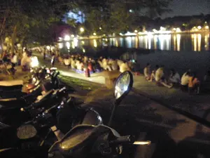 Bức ảnh chụp một khung cảnh về đêm tại một khu vực ven hồ, có khả năng là ở Hà Nội. Rất đông người đang tụ tập ngồi dọc theo bờ hồ trên vỉa hè hoặc bãi cỏ. Họ ngồi trên những chiếc ghế nhựa thấp hoặc trực tiếp trên nền đất để hóng mát, trò chuyện và có thể là uống nước, một hoạt động giải trí phổ biến. Ở phía trước, có một dãy xe máy đang đỗ, trong đó nổi bật là gương chiếu hậu của một chiếc xe ở gần máy ảnh nhất. Phía bên kia hồ, ánh đèn từ đường phố và các tòa nhà phản chiếu lấp lánh trên mặt nước, tạo nên một không gian về đêm rất đặc trưng. Cây cối được trồng dọc theo bờ hồ. Không có bất kỳ biển hiệu hay chữ viết nào có thể nhận dạng được trong ảnh do chất lượng ảnh thấp và chụp vào ban đêm.。（日本語訳）。写真は、おそらくハノイの湖畔エリアの夜景を写したものです。非常に多くの人々が、歩道や芝生の上で湖畔に沿って集まり座っています。彼らは低いプラスチック製の椅子や地面に直接座って、涼んだり、おしゃべりをしたり、そしておそらくは一般的な娯楽活動である飲み物を飲んだりしています。前方にはバイクが一列に駐車しており、その中で最もカメラに近いバイクのバックミラーが目立っています。湖の対岸では、街灯や建物の光が水面にキラキラと反射し、非常に特徴的な夜の空間を作り出しています。湖畔に沿って木々が植えられています。写真の画質が低く、夜間に撮影されたため、写真内には識別できる看板や文字は一切ありません。